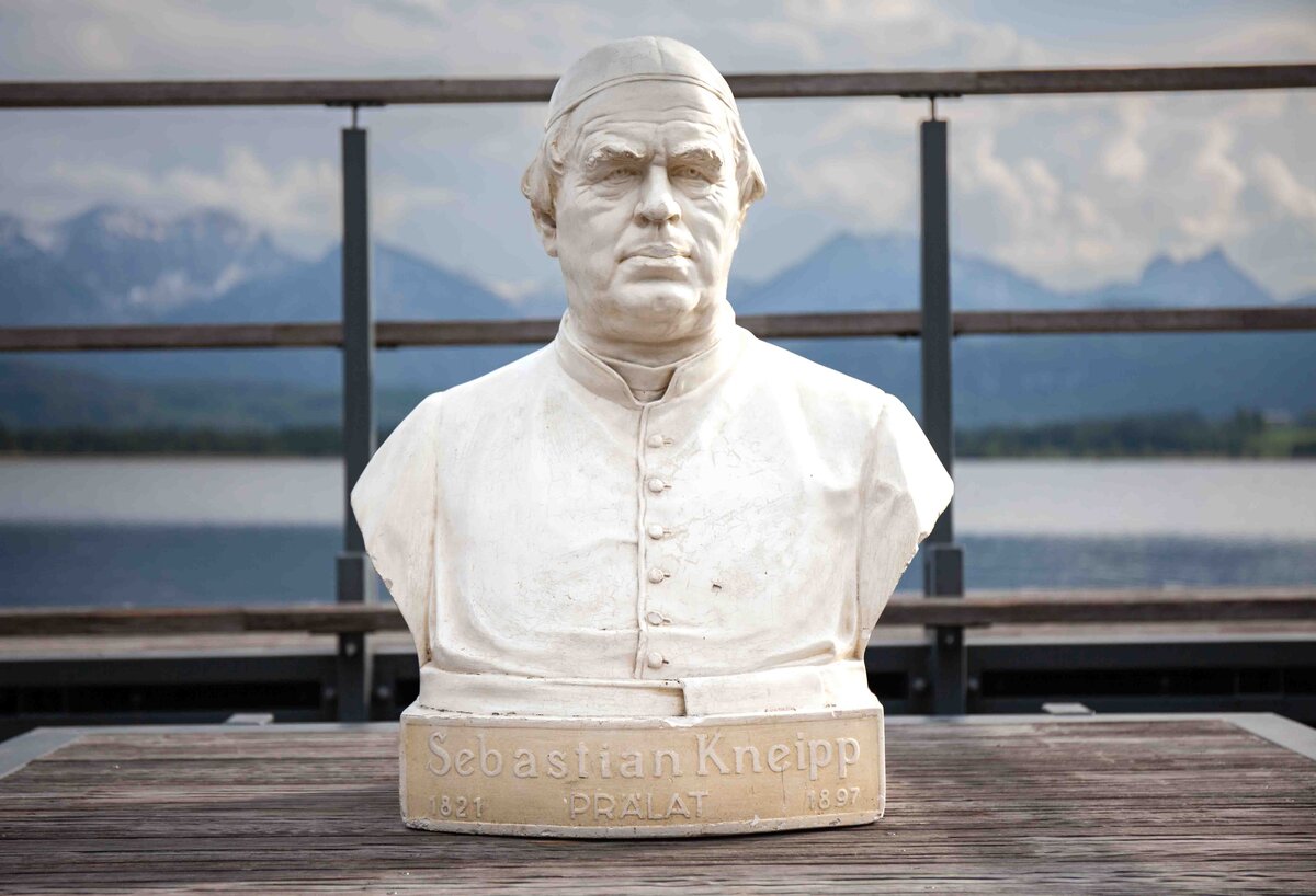 Eine Büste von Pfarrer Sebastian Kneipp, im Hintergrund der Hopfensee und Berge.